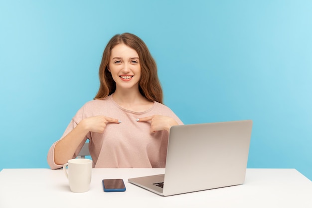 I39m, la mejor empleada feliz y egoísta, señalándose a sí misma y sonriendo orgullosamente alardeando de logros en el trabajo de promoción sentada en el lugar de trabajo con una foto de estudio en el interior de una computadora portátil aislada en un fondo azul