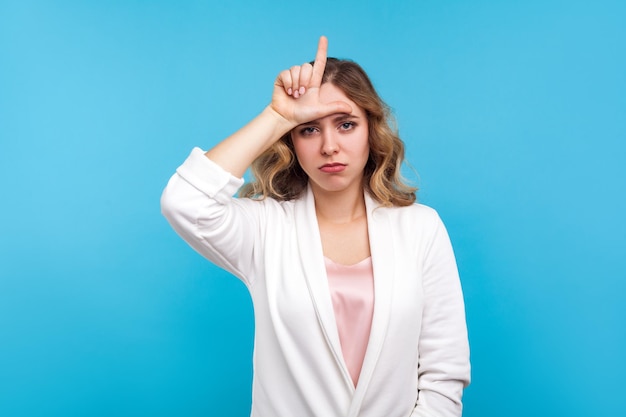 Ich bin ein Verlierer. Porträt einer depressiven Frau mit welligem Haar in weißer Jacke, die Verlierergeste macht, L-Fingerzeichen auf der Stirn und düster aussehend, besorgt über Misserfolge. Studioaufnahme isoliert auf blauem Hintergrund