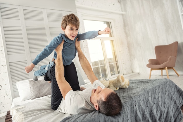 Ich bin inspiriert. Gut aussehender fröhlicher blonder Junge, der lächelt und Spaß mit seinem Vater hat, während er auf dem Rücken auf dem Sofa liegt