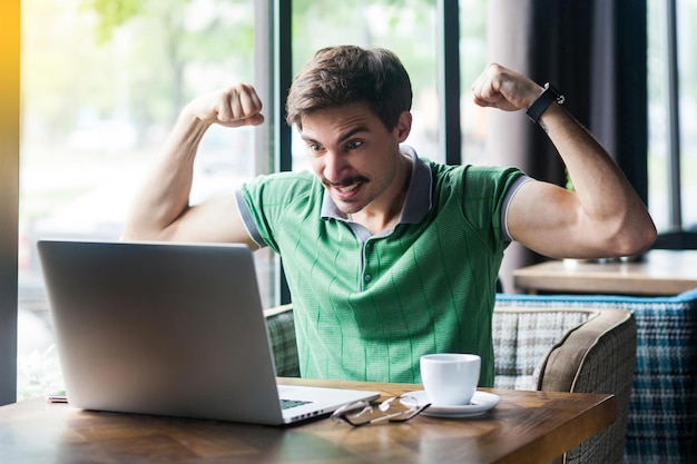 Ich bin stark. Junger aggressiver Geschäftsmann im grünen T-Shirt sitzt, schaut bei Videoanruf auf den Laptop-Bildschirm und zeigt seinen Bizepsmuskel, wütendes Gesicht. Unternehmenskonzept. Innenaufnahme tagsüber in der Nähe des Fensters.