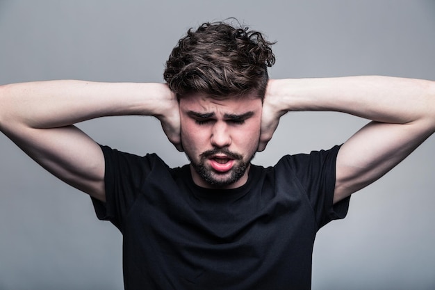 Ich brauche Ruhe Frustrierter junger Mann in Hemd und Krawatte, der die Ohren mit den Händen bedeckt und die Augen geschlossen hält, während er vor grauem Hintergrund steht