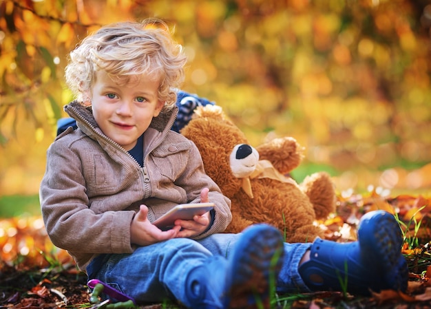 Ich darf überall hin, solange ich mein Telefon habe Zugeschnittenes Porträt eines entzückenden kleinen Jungen, der sein Handy benutzt, während er im Herbst im Freien sitzt