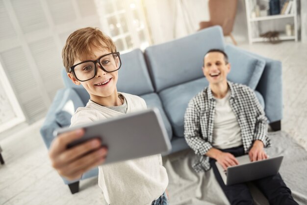 Ich fotografiere gerne. Hübscher freudiger fröhlicher blonder Junge, der lächelt und große Brille trägt und Selfies davon nimmt, während sein Vater im Hintergrund sitzt