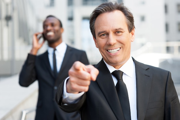 Ich habe Dich ausgewählt! Selbstbewusster älterer Mann in formeller Kleidung, der Sie zeigt und lächelt, während ein afrikanischer Mann im Hintergrund mit dem Handy spricht