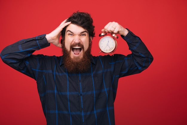 Ich hasse es, zu spät zu sein. Mann halten Wecker in der Hand. bärtiger Mann mit Uhr über roter Wand