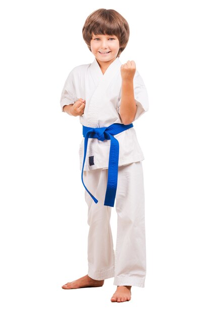 Ich kann mich verteidigen. Volle Länge des kleinen Jungen in Karate-Uniform, der in Karate-Position steht, während er isoliert auf Weiß steht