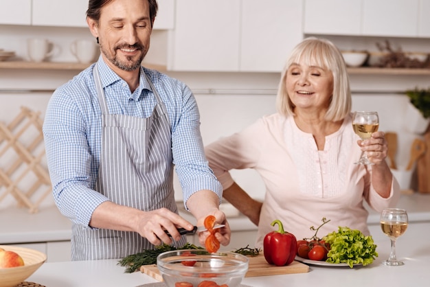 Ich kümmere mich immer um meine Eltern. Erfreuter glücklicher reifer Mann, der in der Küche steht und Tomaten schneidet, während seine ältere Mutter ein Glas Wein genießt