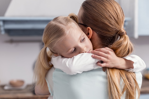 Ich liebe dich. Freundliches schönes kleines Mädchen, das Mutter umarmt, während es beiseite schaut und Pferdeschwänze trägt