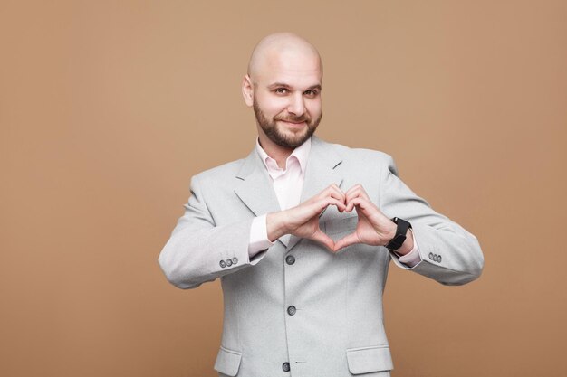 Ich liebe dich. Hoffnungsvoller, gutaussehender, kahlköpfiger Geschäftsmann mittleren Alters im klassischen hellgrauen Anzug, der ein Herzzeichen zeigt und mit einem Lächeln in die Kamera schaut. Indoor-Studioaufnahme, auf braunem Hintergrund isoliert