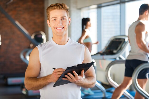 Ich mache dich gesund! Schöner junger Mann, der die Zwischenablage hält und lächelt, während die Leute im Hintergrund auf dem Laufband laufen