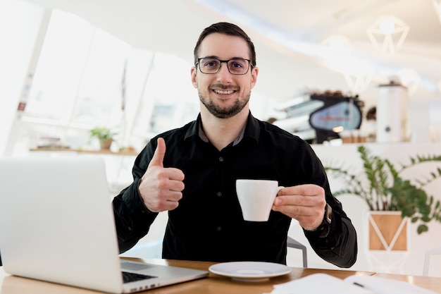 Ich mag diesen Kaffee sehr! Attraktiver Mann mit Brille und schwarzem Hemd, das eine Tasse flaches Weiß hält, lächelt und nach oben blättert. Helles Café-Interieur im Hintergrund. Kaffee hilft dir, dich zu konzentrieren.