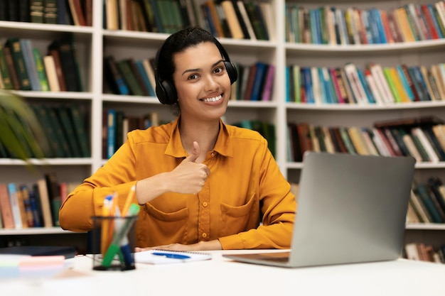 Ich mag Fernlernen, glückliche Studentin, die Daumen hoch gestikuliert, sitzt am Schreibtisch mit Laptop drin