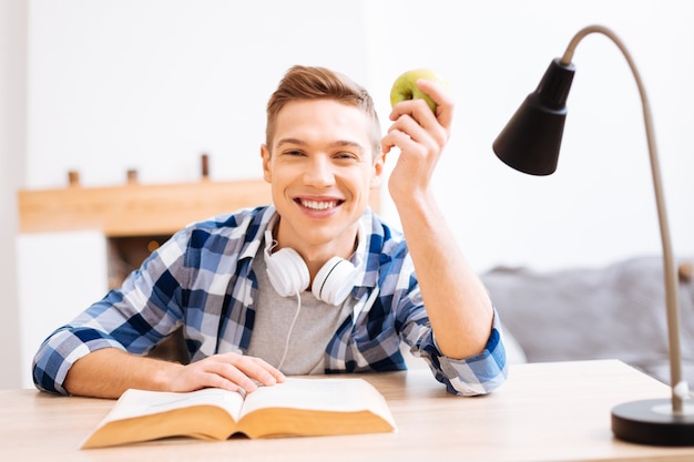 Foto ich mag früchte. gut aussehender entzückter blonder junge, der lächelt und mit einem buch am tisch sitzt und einen apfel hält