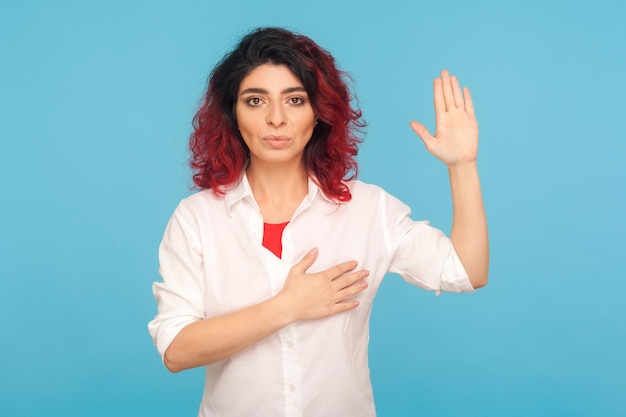 Ich schwöre. Porträt einer Hipster-Frau mit ausgefallenen roten Haaren, die die Hand auf dem Herzen hält und die Handfläche erhebt, um einen Eid zu leisten, sich hingibt und verspricht, die Aufgabe verantwortungsbewusst zu erfüllen. Studioaufnahme im Innenbereich, blauer Hintergrund