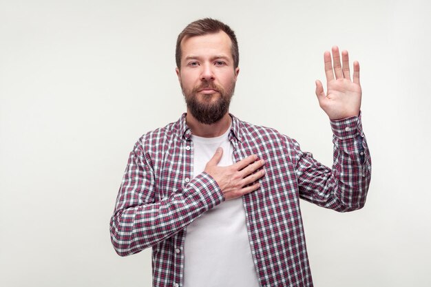 Ich schwöre Porträt eines verantwortungsbewussten bärtigen Mannes in kariertem Hemd, der die Hand auf der Brust hält und verspricht, die Wahrheit zu sagen, ehrlich zu sein, engagierte Innenstudioaufnahme isoliert auf weißem Hintergrund