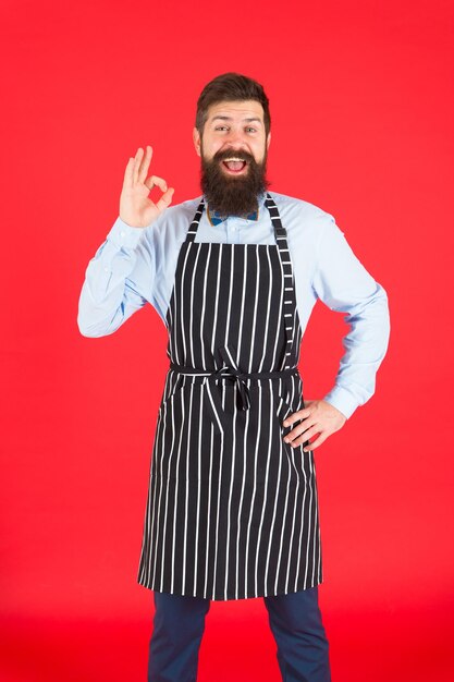 Ich weiß alles über das Kochen. Koch des bärtigen Mannes. Hipster, der zu Hause oder im Restaurant kocht. Modernes Café-Konzept. Kochen moderner Gerichte. Chefkoch roter Hintergrund. Mann mit Bartkoch-Hipster-Schürze.