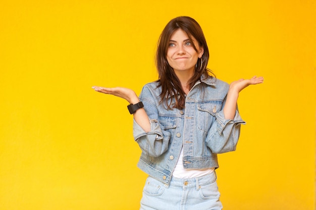 Ich weiß nicht. Porträt einer verwirrten, schönen, brünetten jungen Frau mit Make-up im lässigen Denim-Stil, die mit erhobenen Armen steht, wegschaut und zweifelt. Indoor-Studioaufnahme, isoliert auf gelbem Hintergrund