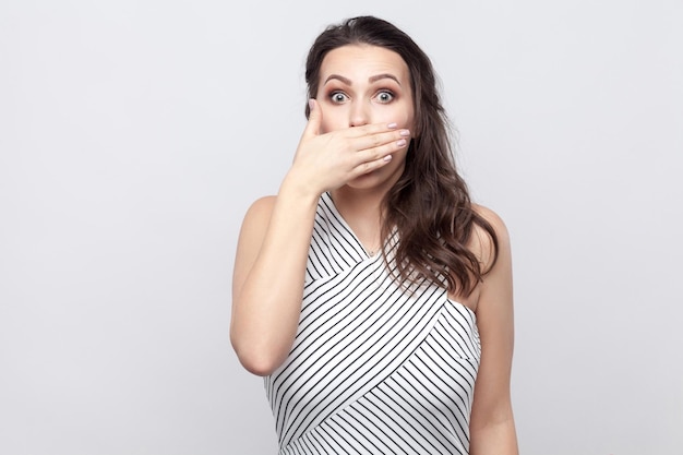 Ich werde schweigen. Porträt einer schockierten schönen jungen brünetten Frau mit Make-up und gestreiftem Kleid, die mit großen Augen steht und ihren Mund bedeckt. Indoor-Studioaufnahme, isoliert auf grauem Hintergrund.