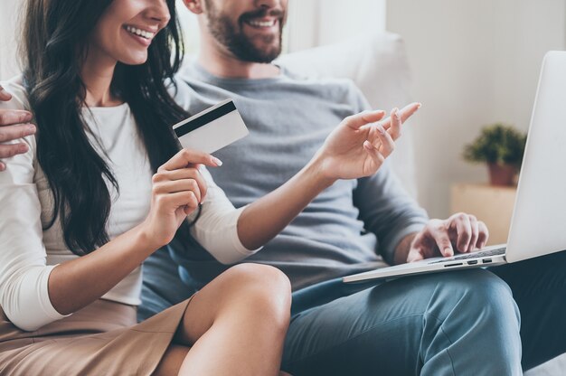 Ich will das! Nahaufnahme einer schönen jungen Frau, die Kreditkarte hält und mit einem Lächeln auf den Laptop zeigt, während sie zusammen mit ihrem Mann auf der Couch sitzt