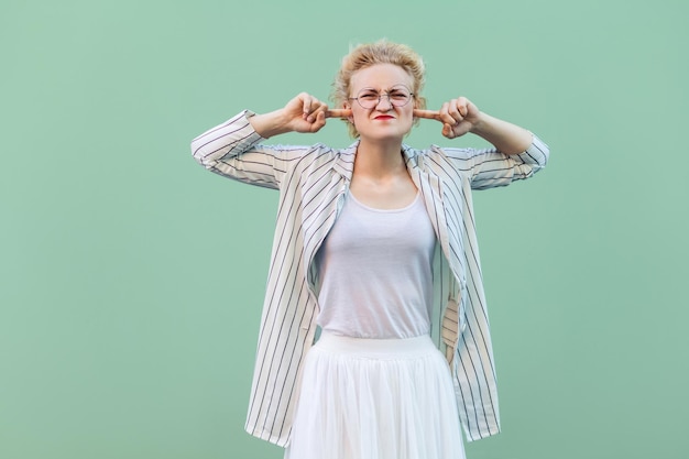 Ich will das nicht hören. Junge Frau in weißem Hemd, Rock, gestreifter Bluse mit Brille, die mit geschlossenen Augen und Finger an den Ohren steht. Indoor-Studioaufnahme auf hellgrünem Hintergrund isoliert.