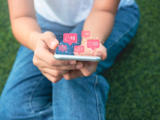 Foto Ícone de mídia social moderna em balão rosa flutuando em torno do smartphone nas mãos de uma mulher sentada na grama verde com gesto de relaxamento, estilo de vida, tecnologia e conceito de rede social.