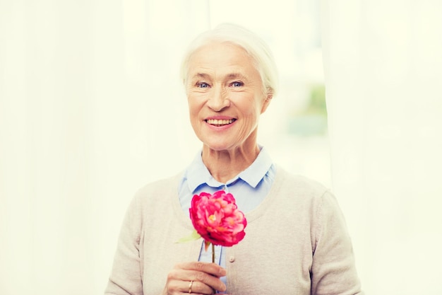 idade, feriados e conceito de pessoas - feliz sorridente mulher sênior com flor em casa