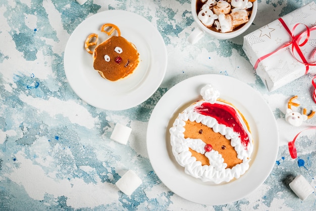 Foto idea para un desayuno de navidad para niños: panqueques decorados como santa claus y ciervos, cacao con ositos de peluche y malvaviscos de ciervos. sobre una superficie clara, con espacio de copia de vista superior de regalo de navidad