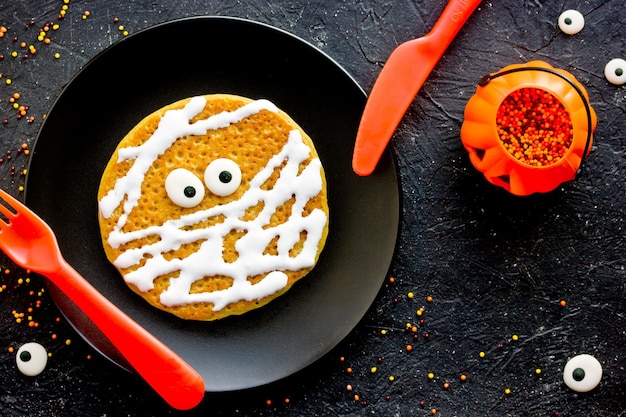 Idea divertida de panqueque de momia para los niños para el desayuno de Halloween panqueque de calabaza con crema y ojos de caramelo en forma de momia en la vista superior de la placa negra