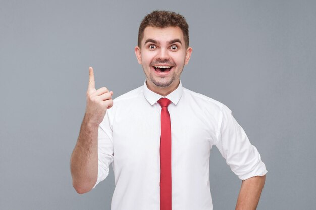 Idea Retrato de un joven apuesto hombre sorprendido con camisa blanca y corbata de pie y mirando a la cámara con gesto de idea y disfrutar de interiores aislados en fondo gris