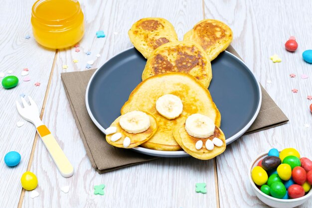 Ideia de comida de almoço de café da manhã saudável criativa engraçada de páscoa para crianças coelhinho feito de panquecasbanana com doces de melovos no fundo da mesa de madeira de pratoVista superior Postura plana