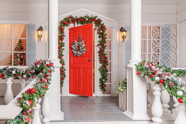 Ideia de decoração de varanda de Natal. Entrada da casa com porta vermelha decorada para férias. Guirlanda de grinalda vermelha e verde de galhos de árvores de abeto e luzes na grade. Véspera de Natal em casa.