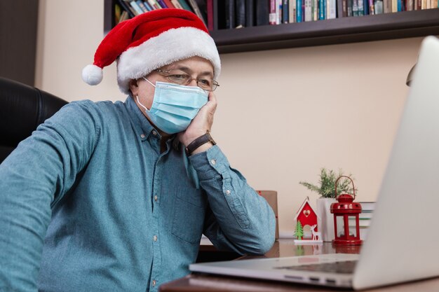 Ideia de parabéns online de Natal. Último homem com chapéu de Papai Noel fala usando laptop para amigos de videochamada e crianças. O quarto está decorado de forma festiva. Natal durante o coronavírus.