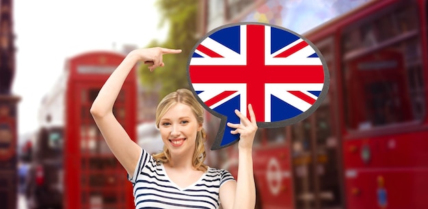 idioma extranjero, inglés, viajes, personas y concepto de comunicación - mujer sonriente sosteniendo una burbuja de texto de bandera británica y señalando con el dedo el fondo de la calle de la ciudad de Londres