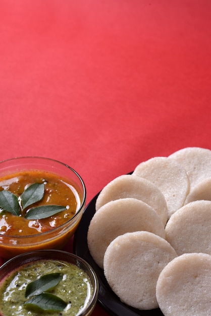 Idli mit Sambar- und Kokos-Chutney auf roter Oberfläche, indisches Gericht: südindisches Lieblingsessen Rava Idli oder Grieß untätig oder Rava untätig, serviert mit Sambar und grünem Chutney.