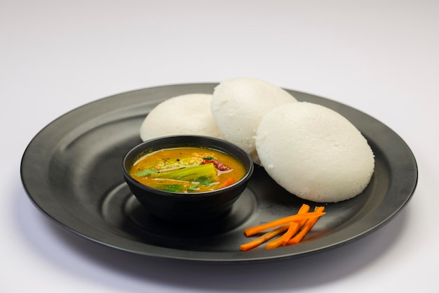 Idly o Idli, artículo principal del desayuno del sur de la India, que está bellamente dispuesto en un plato negro sobre fondo blanco.