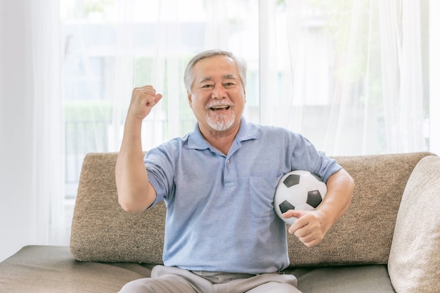 Idoso animado, homem idoso se sentindo feliz segurando uma bola de futebol de futebol, prepare-se para a torcida favorita do time no sofá em casa, estilo de vida asiático idoso conceito de boa saúde