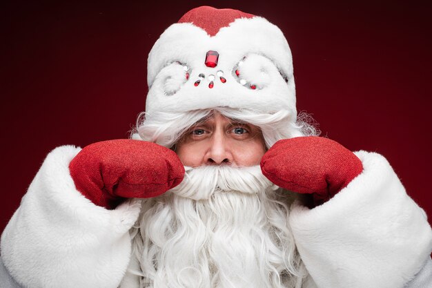 Idoso bigode grisalho barbudo homem de Papai Noel com chapéu de Natal posando isolado na parede vermelha.