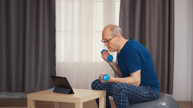 Idoso com idade masculina exercitando-se com halteres sentado na bola suíça durante o treinamento online. Idoso reformado treino saudável saúde desporto em casa, exercício de actividade física na velhice