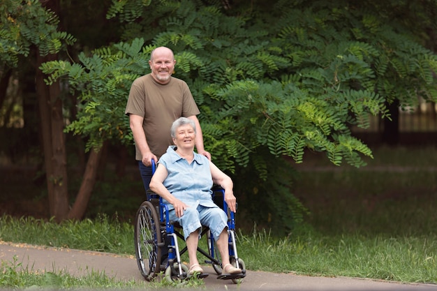 Idoso feliz caminhando com uma senhora idosa com deficiência sentada em uma cadeira de rodas ao ar livre