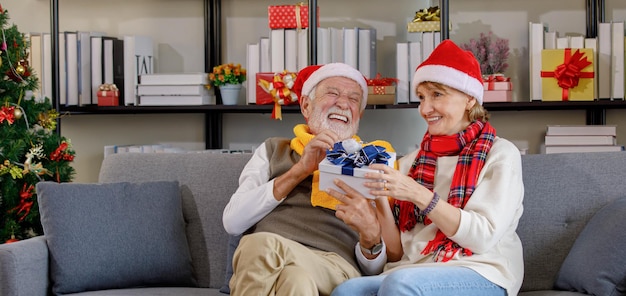 Idoso feliz rindo enquanto está sentado no sofá e recebendo um presente da esposa madura com um lenço e um chapéu de Papai Noel durante a celebração do Natal em casa