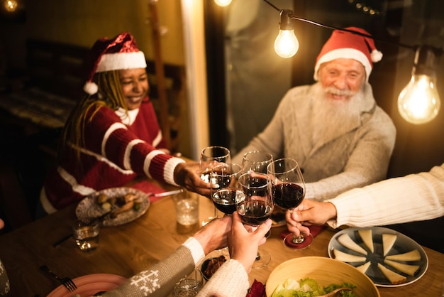 Idosos felizes torcendo por vinho durante o jantar de Natal em casa usando chapéus de Papai Noel - Foco nos copos