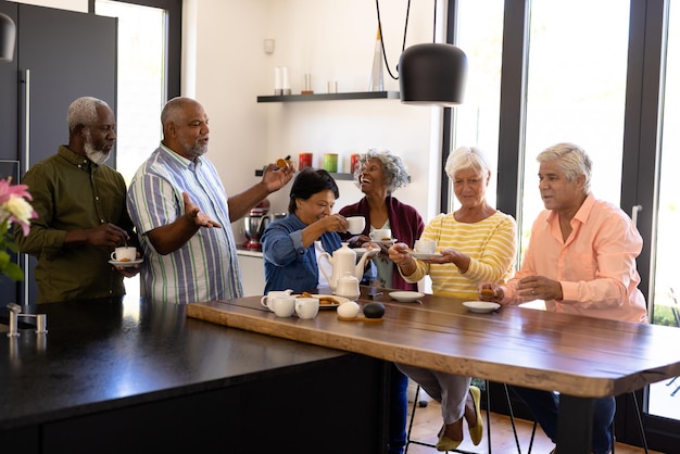 Foto idosos multirraciais alegres conversando e desfrutando de café, biscoitos na mesa de madeira em casa de repouso. lanches, bebidas, comida, inalterados, amizade, união, apoio, vida assistida e aposentadoria.