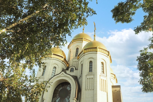 Iglesia ortodoxa de ladrillo con cúpulas doradas en el contexto de un hermoso cielo nublado y abedules.