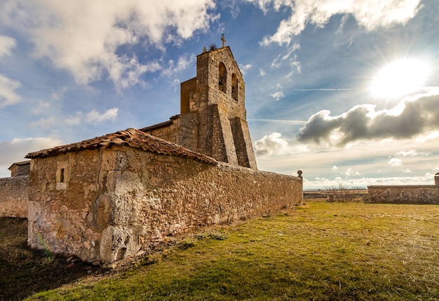 Igreja de pedra muito antiga no meio do campo com céu azul, nuvens e sol. Meio rural. Segovia