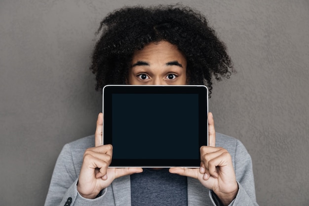 Ihre Lösung ist hier. Junger afrikanischer Mann, der sein Gesicht hinter digitalem Tablet mit Kopienraum versteckt, während er vor grauem Hintergrund steht