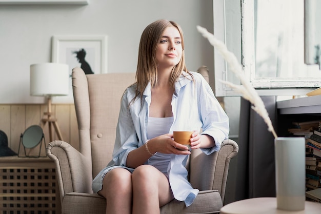 Ihren frischen Kaffee genießen Schöne junge Frau, die Kaffee trinkt und durch das Fenster schaut, während sie zu Hause im Sessel sitzt