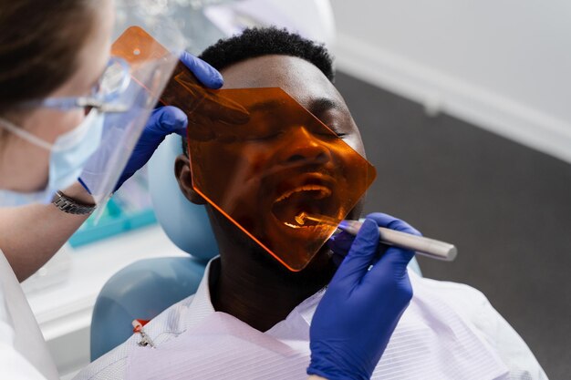 Iluminación ultravioleta del relleno dental de fotopolímero para el hombre africano en odontología El dentista en gafas protectoras trata y elimina la caries del paciente africano