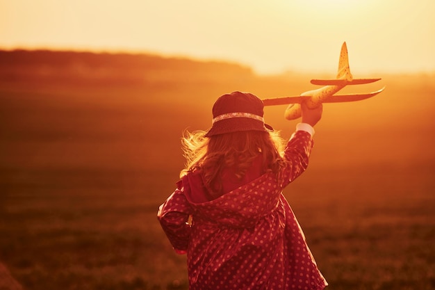 Iluminada por la luz del sol de color naranja Una niña linda se divierte con un avión de juguete en el hermoso campo durante el día