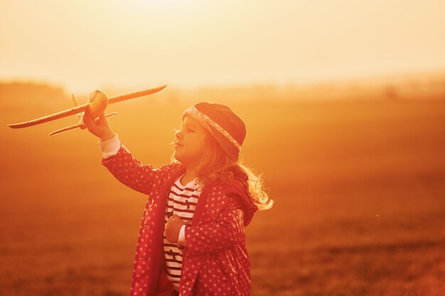 Iluminado por la luz solar de color naranja. Niña linda divertirse con avión de juguete en el hermoso campo durante el día