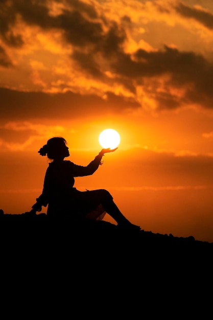 Foto ilusão óptica de silhueta de mulher segurando o sol contra o céu dramático laranja durante o pôr do sol